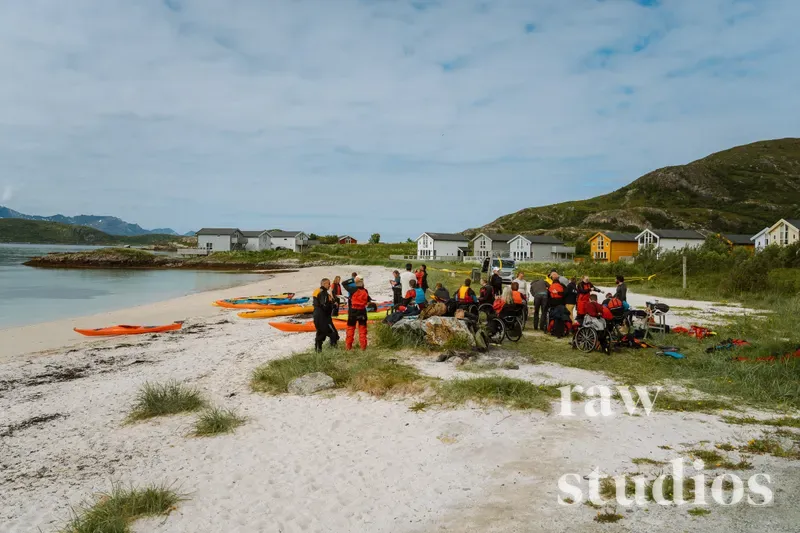 En gruppe mennesker i fargerike padlevester samlet ved oransje og gule kajakker på en lys sandstrand ved kysten. Under en blå sommerhimmel med fjell og små hus i bakgrunnen er stemningen preget av lyst dagslys og aktivt friluftsliv.
