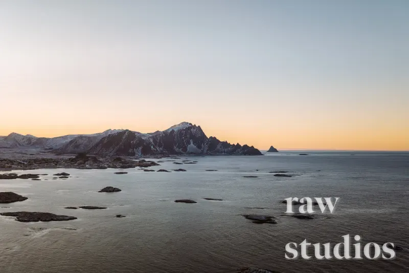 Snødekte fjell og klipper ved havet under en arktisk solnedgang. Kystlandskapet viser et rolig vann i blåtoner mot en varm, lysende horisont i vintertiden.