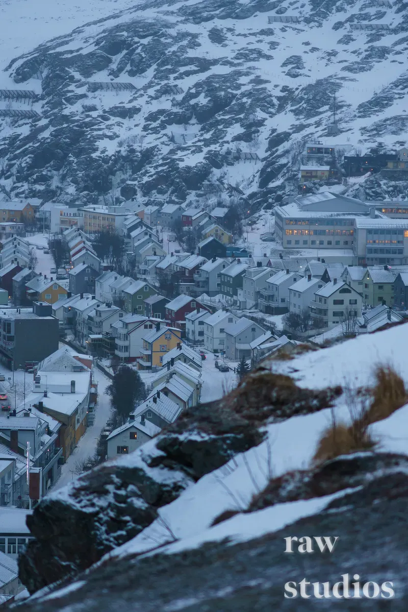 Snødekt kystby med fargerike hus ved foten av et bratt og steinete fjell. Landskapet preges av vinter, kalde toner og en rolig stemning i det snølagte terrenget.