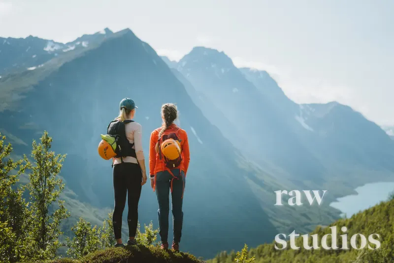 To kvinner med ryggsekker og hjelmer står i et fjellandskap og ser utover en dal med vann og skog. Scenen viser grønne omgivelser og høye fjelltopper under en lys sommerhimmel, preget av en stemning av friluftsliv og eventyr.
