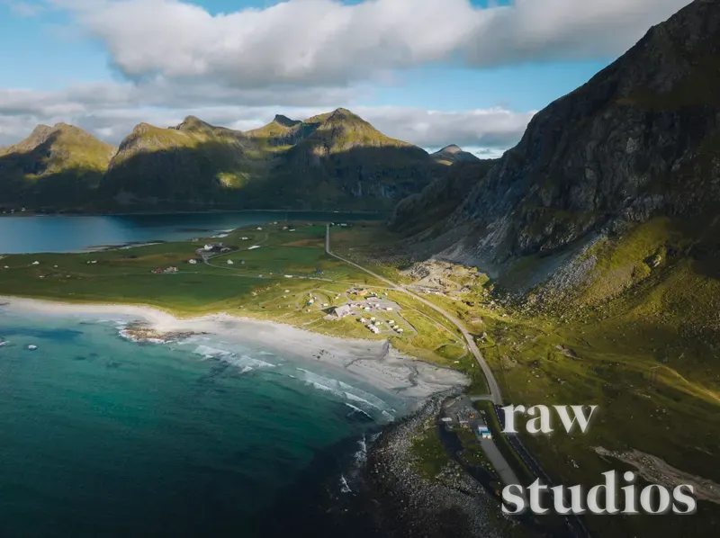 Et dronebilde av et kystlandskap med dramatiske fjell, en vei langs havet og små hus ved en sandstrand. Bildet viser grønne marker og blått vann i sommerlig dagslys under en delvis skyet himmel.