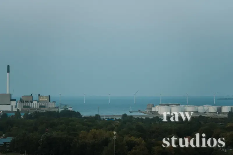 Industrianlegg ved kysten med lagertanker, fabrikker og vindturbiner i det fjerne over havet. Forgrunnen består av grønn skog under en overskyet, blågrå himmel. Bildet har en rolig og dempet stemning.