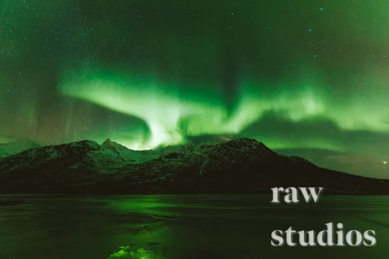Grønt nordlys over snødekte fjell og et mørkt vann under en stjerneklar nattehimmel. Vinterlandskapet er preget av magisk lys og en rolig stemning.