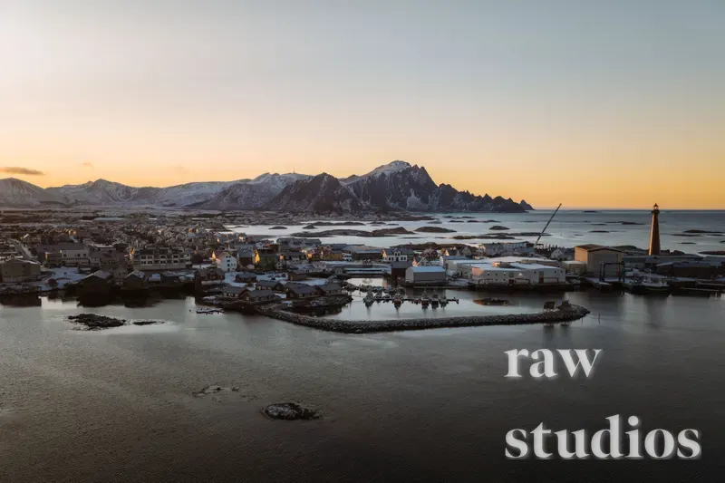 Droneutsikt over en kystby med havn ved foten av snødekte fjell under en solnedgang. Det gylne lyset fra skumringen reflekteres i sjøen og det vinterlige landskapet.