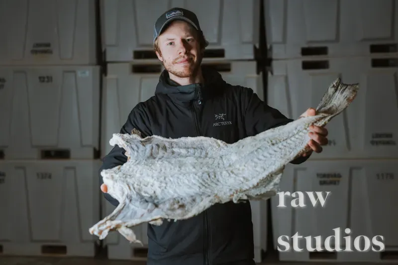 En mann holder opp et stort stykke tørrfisk foran stablede hvite plastkasser i et lager eller produksjonslokale. Bildet har dempet belysning og nøytrale farger i et innendørs miljø knyttet til fiskeri eller havbruk.