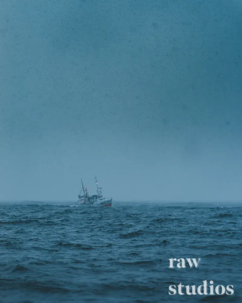 En liten båt seiler på det urolige havet under en grå, overskyet himmel. Det blågrå vannet med bølger skaper et rått og stemningsfullt maritimt landskap.