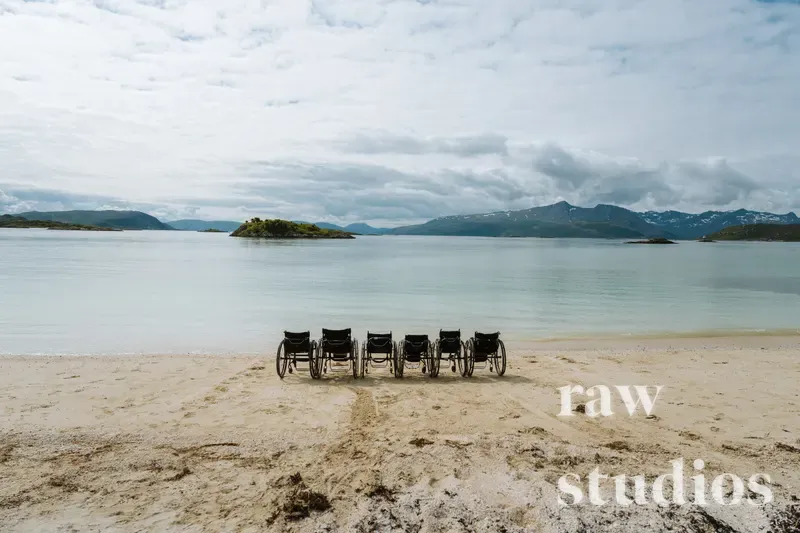 Seks svarte strandstoler står på rekke langs en sandstrand ved et rolig hav og kystlandskap med fjell og øyer i bakgrunnen. Bildet har dempede farger i grått, blått og beige under en overskyet himmel, noe som skaper en fredelig stemning.