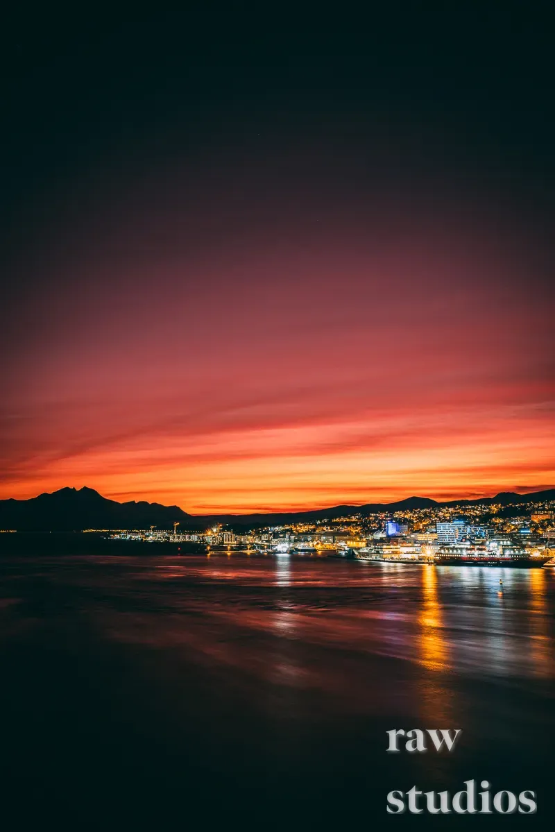 Et panorama over en kystby ved skumringstiden med en brennende oransje og lilla himmel. Lys fra bebyggelse og havn reflekteres i sjøen foran mørke fjellsilhuetter.