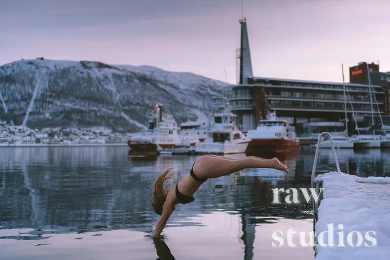 En person i mørk badedrakt dykker mot vannet i en snødekt havn med båter og fjell i bakgrunnen. Scenen viser vinterlig aktivitet ved kysten under et dempet lys.