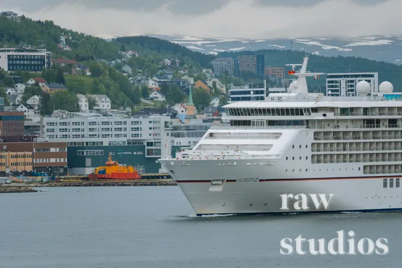 Et stort hvitt cruiseskip seiler gjennom en fjord ved en kystby med bebyggelse i skråningen og snødekte fjell i bakgrunnen. Bildet viser et maritimt landskap med rolig vann, grønne åser og overskyet dagslys.