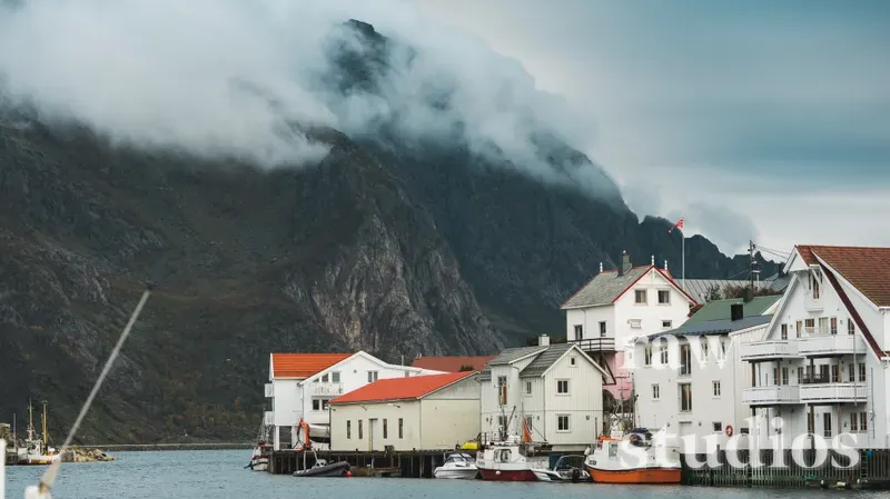 Hvite hus med røde tak ligger ved kysten i en havn med små båter fortøyd i sjøen. Dramatiske fjell dekket av lavt skydekke rager over bebyggelsen under en grå og stemningsfull himmel.