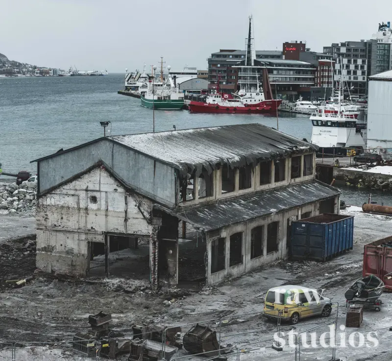 En slitt og delvis ødelagt industribygning ligger ved en havn med skip, båter og bybebyggelse i bakgrunnen. Det maritime miljøet er preget av dempede farger under en grå og overskyet himmel.