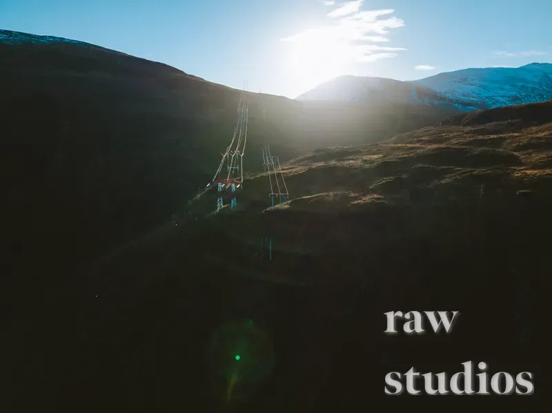 Dronebilde av kraftlinjer som strekker seg gjennom et mørkt fjellandskap med snødekte topper under en lysende sol. Motivet viser infrastruktur i et øde landskap preget av sterke kontraster mellom lys og skygge.