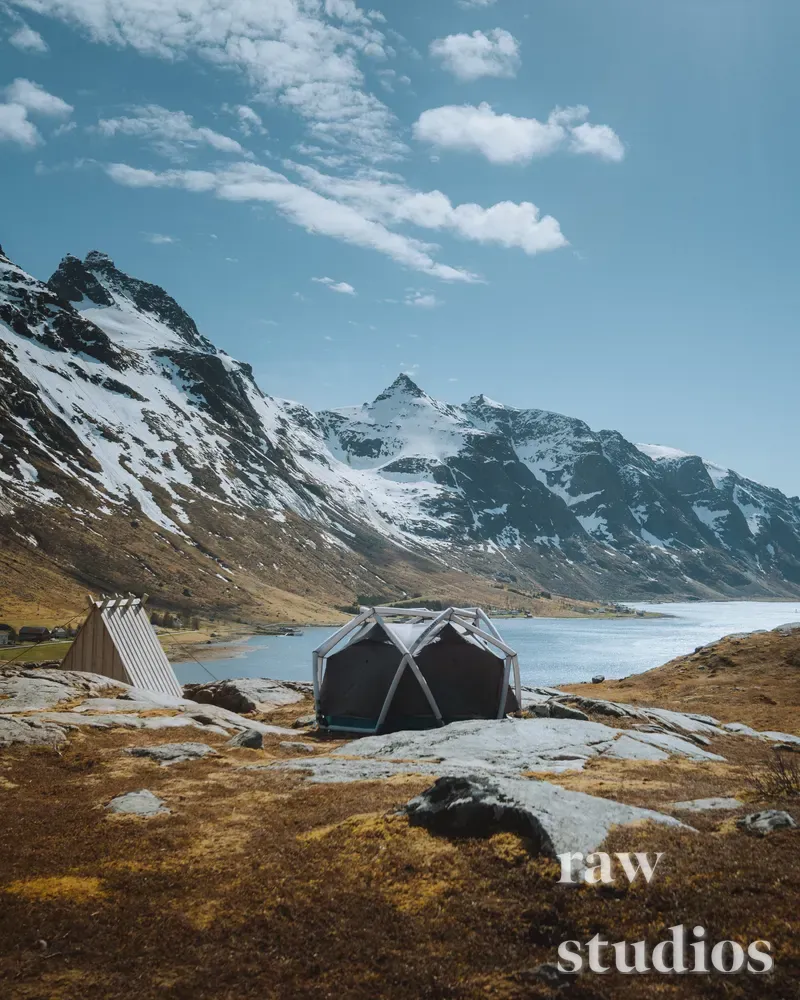 Et mørkegrått telt og en liten trekonstruksjon står på en steinete fjellhylle ved siden av en blå innsjø under snødekte fjelltopper. Landskapet preges av blå himmel, hvitt snø og gylne toner i terrenget under klart dagslys med en rolig stemning fra camping i naturen.