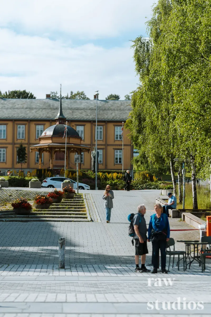 Folk vandrer på en solfylt plass omgitt av grønne trær og blomstrende krukker i et sommerlig bymiljø. I bakgrunnen står en stor, gul trebygning med et karakteristisk tårn under en lys himmel. Scenen er preget av dagslys og en rolig atmosfære.