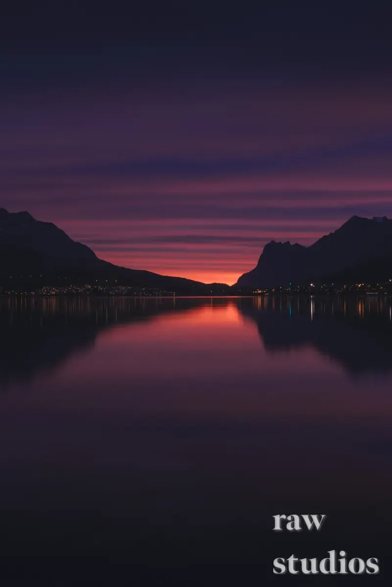 Et landskap med mørke fjell som speiler seg i en stille innsjø under en lilla og oransje solnedgang. Stemningen er rolig og fredelig i skumringen.