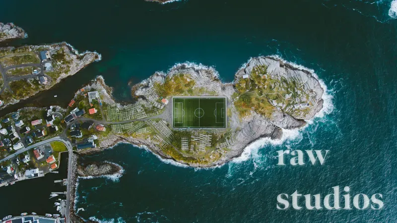 Et dronebilde viser en grønn fotballbane på en liten øy eller holme omgitt av mørkeblått hav og bølger som slår mot land. Kystlandskapet består av klipper, vann og enkelte bygninger under klart dagslys.