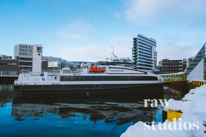 En hvit og svart passasjerbåt ligger ved en kai i en snødekt byhavn under en klar blå himmel. Det stille vannet reflekterer båten og de omkringliggende bygningene i et rolig vinterlandskap.