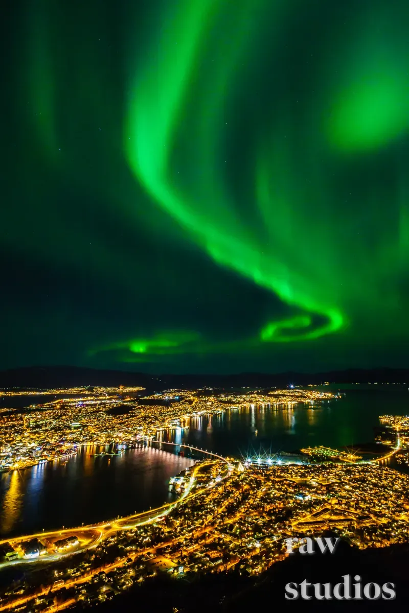 Et nattlig panorama av en opplyst kystby under et grønt nordlys som bølger over himmelen. Det mørke vannet reflekterer lysene fra byen og de strålende aurora-formasjonene i en stemningsfull nattehimmel.