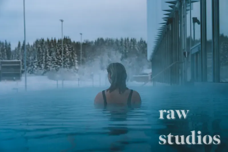 En person står med ryggen til i et dampende utendørsbasseng omgitt av snødekt skog og en moderne glassbygning. Bildet har kjølige blåtoner og viser bading i varmt vann under vinterforhold.