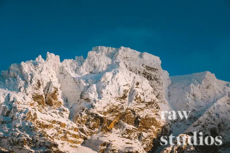 Snødekte fjelltopper og bratte fjellsider under en klar, blå himmel i et vinterlandskap. Kontrasten mellom hvit snø og mørke klipper skaper en kald og rolig stemning i det øde fjellet.
