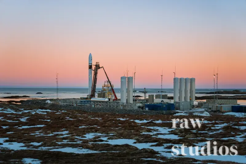 En rakett står på en utskytingsrampe i et snødekt kystlandskap ved solnedgang. Ved havet ser man industriell infrastruktur under en rosa og blå himmel i arktisk vinterlys.