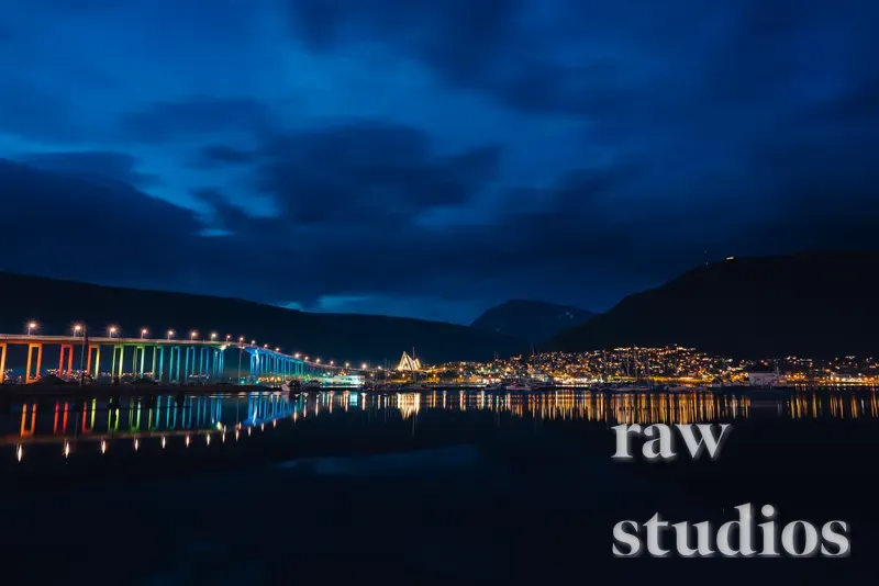 Et nattlig landskap med en opplyst bro i regnbuens farger over en rolig fjord eller kyst. Mørke fjell og en lysende by rammer inn motivet under en blå himmel, med tydelige refleksjoner i vannflaten.