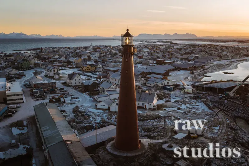Et dronebilde av et høyt fyrtårn som rager over en snødekt kystlandsby ved havet under en solnedgang. Landskapet viser fjell og vann i varme farger mot vinterens kalde omgivelser langs sjøen.