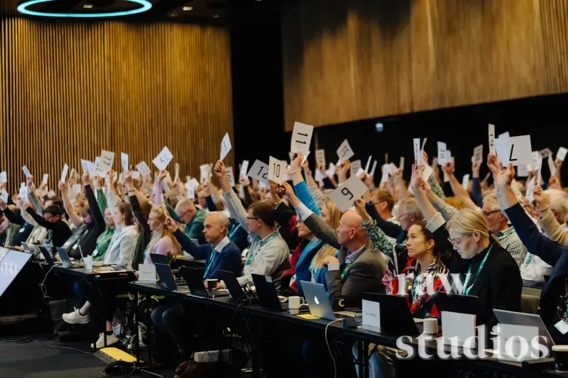 En gruppe mennesker sitter ved bord med bærbare datamaskiner i et konferanselokale og holder opp hvite kort med tall under et arrangement. Lokalet har trevegger og varm belysning som skaper en aktiv stemning.