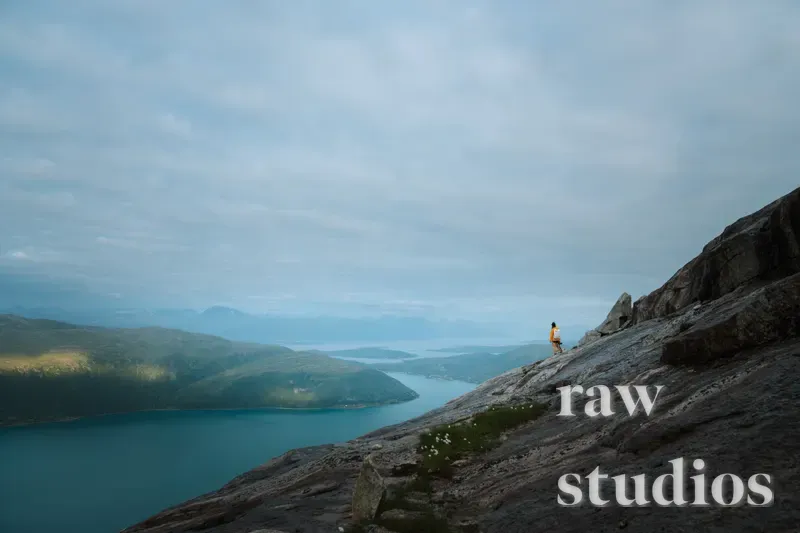 En person står på en steinete fjellrygg med utsikt over en fjord og et vidstrakt kystlandskap under en overskyet himmel. Bildet har dempede blå- og gråtoner og formidler en rolig stemning i et sommerlig fjellmiljø.