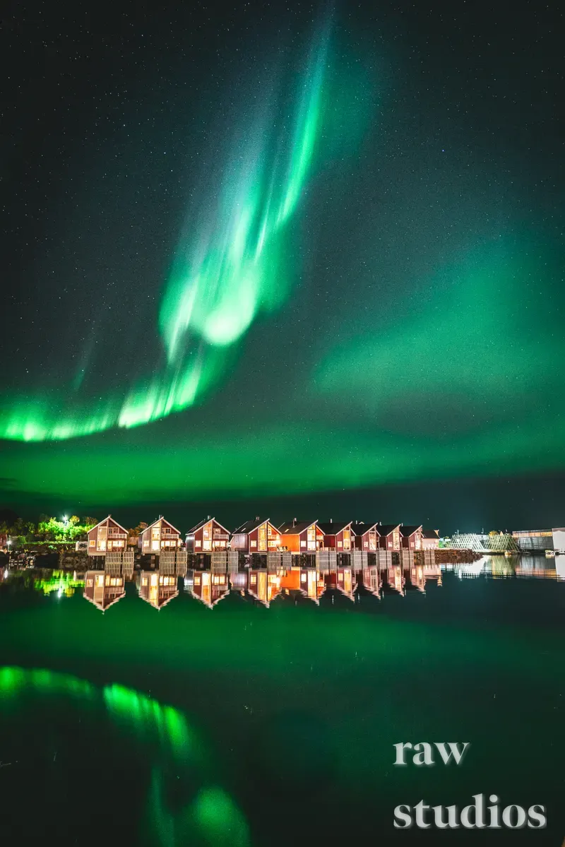 Grønt nordlys over en rekke opplyste rorbuer ved kysten i nattemørket. De varme lysene fra husene speiles i det stille vannet under en stjerneklar himmel.