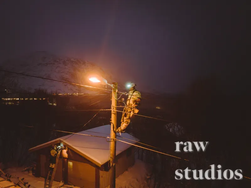 En person arbeider ved en strømmast i et mørkt, snødekt fjellandskap om natten. Varmt lys fra en lykt lyser opp infrastrukturen mot den kalde vinternatten.