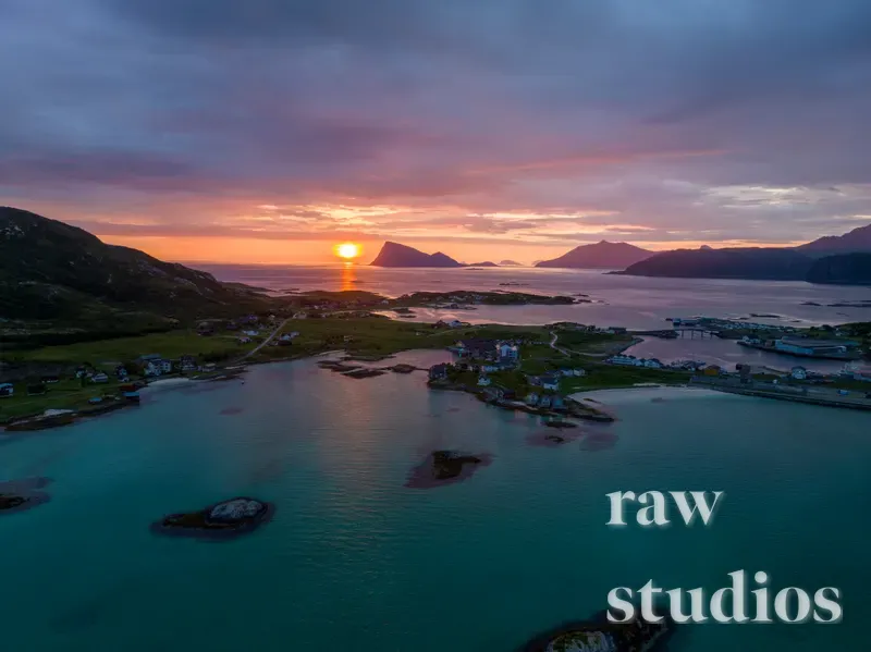 Dronebilde av en solnedgang over et kystlandskap med fjell, øyer og små hus ved havet. Himmelen gløder i oransje og rosa toner som reflekteres i det blå vannet og fjorden. Stemningen er rolig og fredelig i dette åpne landskapet.