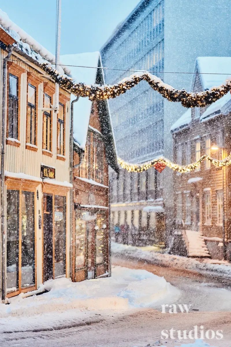 Snødekt bygate med historiske trehus og en moderne bygning under et lett snøfall. Lysende juleguirlander over gaten skaper en varm og stemningsfull atmosfære i vinterlandskapet.