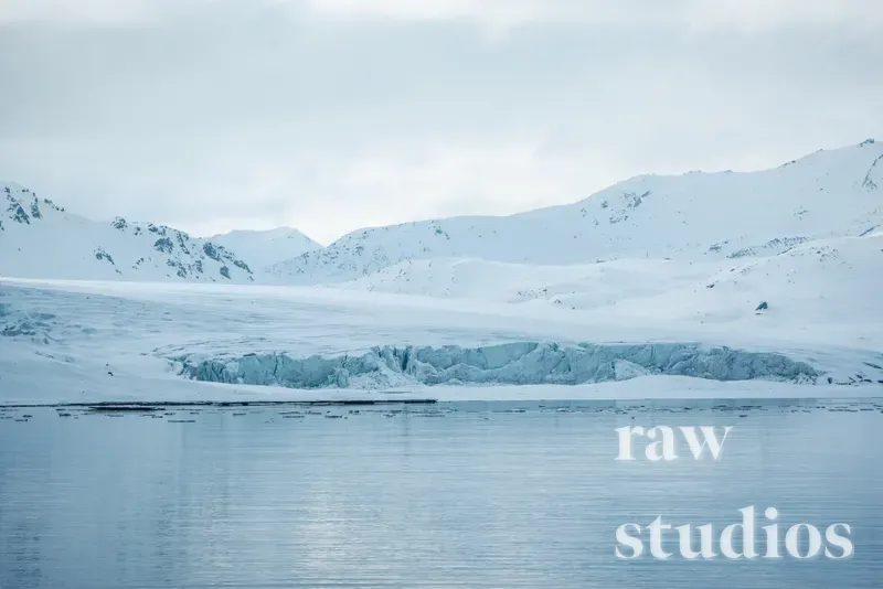 Et arktisk landskap med en isbre som møter havet mellom snødekte fjell. Motivet er preget av kalde farger i blått og hvitt under en overskyet himmel, noe som skaper en rolig vinterstemning.