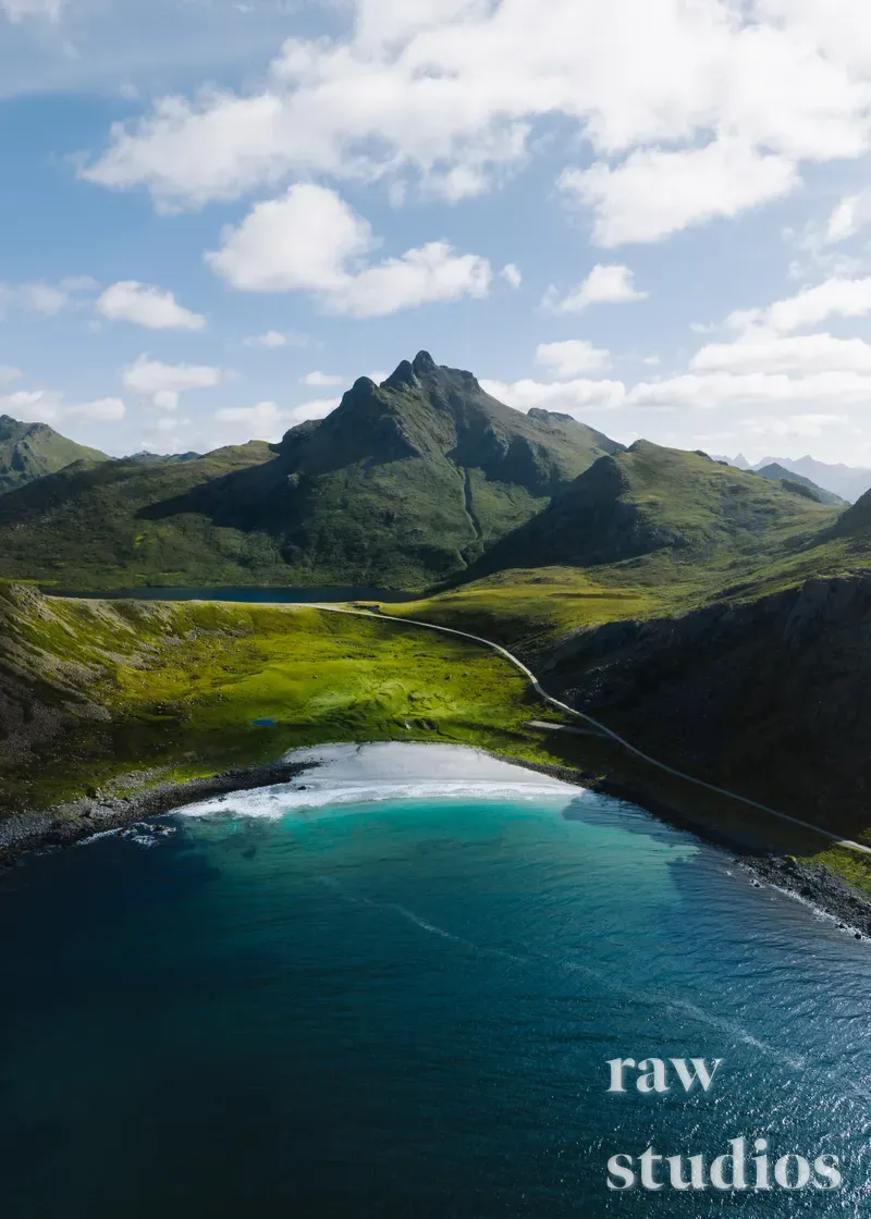 Luftfoto av et kystlandskap med grønne fjellsider, en vei og en strand ved et turkis hav. Bildet viser et sommerlig landskap under en lett skyet himmel med klart dagslys.