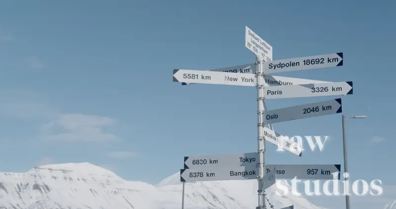 Et skilt med mange retningspiler mot ulike byer står i et vinterlig fjellandskap med snødekte topper under en klar, blå himmel.