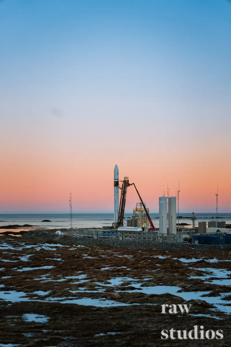 En rakett står på en utskytingsrampe i et snødekt kystlandskap under en fargerik soloppgang eller solnedgang. Motivet viser industriell infrastruktur ved havet med nyanser av oransje og blått i det arktiske vinterlyset.