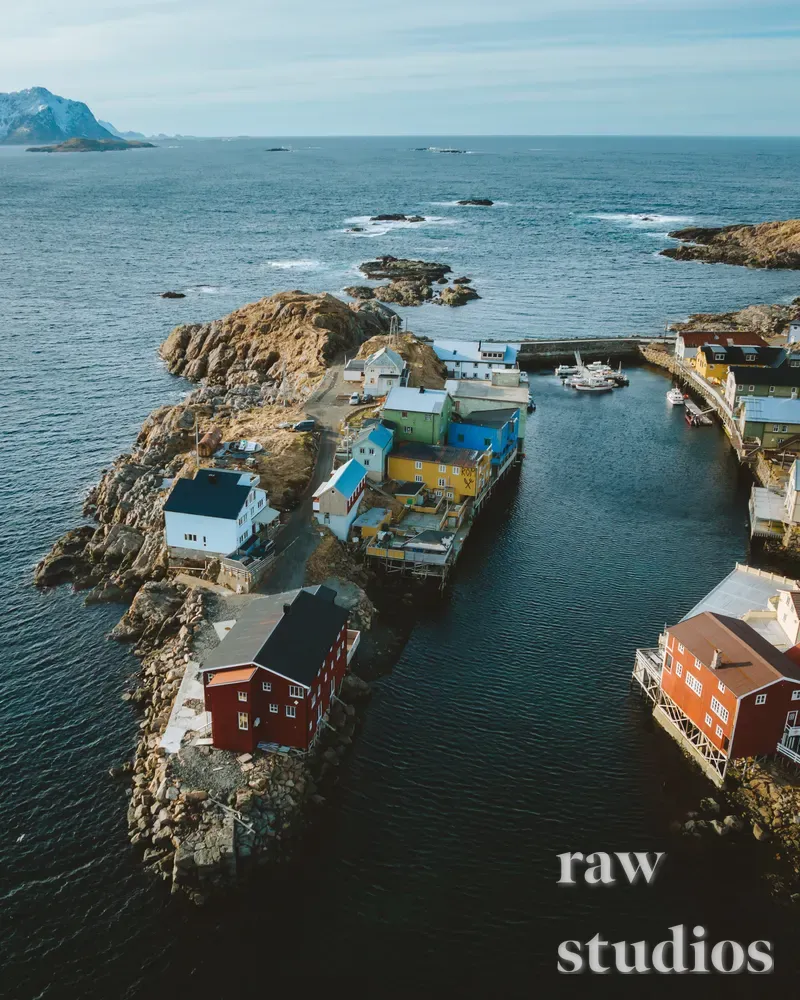 Luftfoto av en maritim kystlandsby med fargerike hus plassert på svaberg og brygger ved havet. Blått vann omgir de mørke klippene, og små båter ligger i den rolige havnen under sommerlig dagslys.