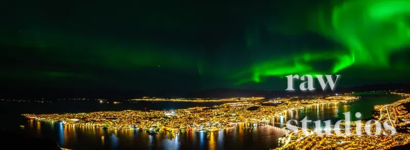 Grønt nordlys over en opplyst kystby ved havet under en mørk nattestimmel. Landskapet består av fjell og vann med varme lys fra bebyggelsen i vintermørket.