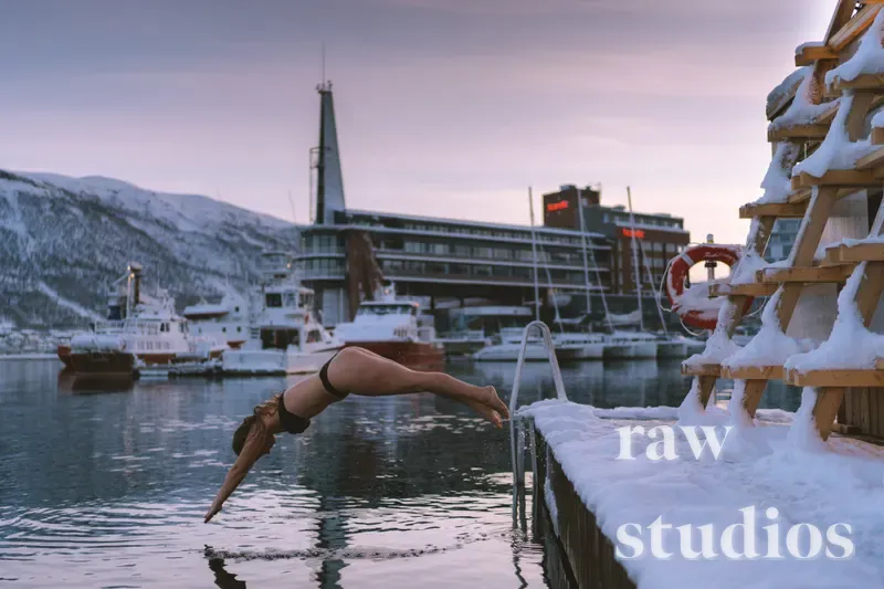 En person dykker ned i sjøen i en snødekt havn med fjell og båter i bakgrunnen. Bildet viser en trebrygge under en stemningsfull skumring med kalde vinterfarger og lys fra solnedgangen.