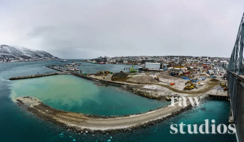 Et dronebilde viser et anleggsområde ved kysten med snødekte fjell under en overskyet himmel. I havnen pågår det utfylling av landmasse med anleggsmaskiner omgitt av blått vann og jordfarger.
