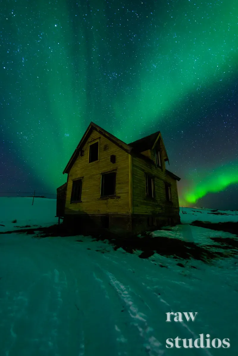 Et gammelt trehus står i et snødekt landskap under et grønt nordlys på en stjerneklar nattehimmel. Scenen viser et arktisk vinterlandskap med kaldt lys og en stille stemning.