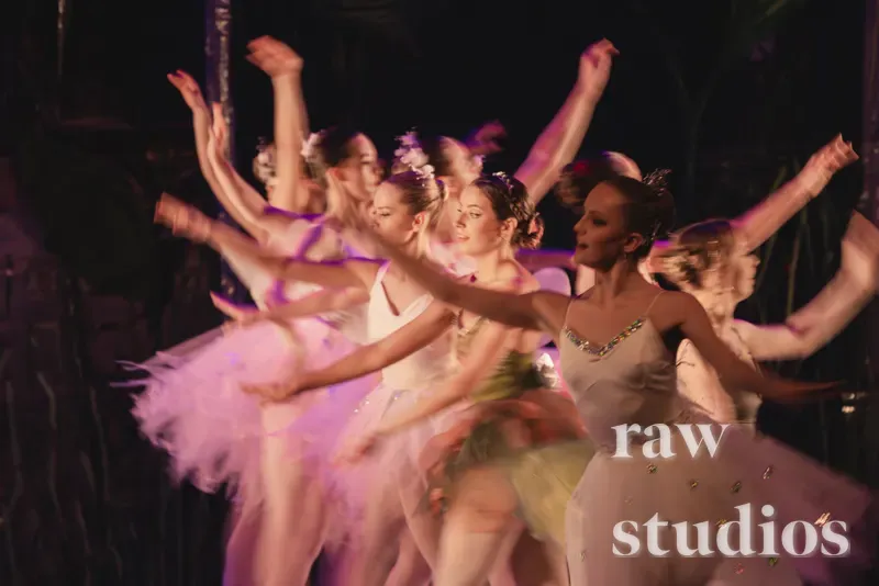 En gruppe ballettdansere i rosa og hvite tutuer utfører en dans på en mørk scene under dramatisk belysning. Scenen preges av bevegelse og en elegant stemning med fokus på de lyse draktene mot den skyggelagte bakgrunnen.
