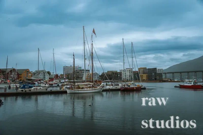 Flere seilbåter og motorbåter ligger fortøyd i en rolig havn med bygninger og fjell i bakgrunnen. Det er en overskyet dag med blåtoner i vannet og himmelen, noe som skaper en fredelig maritim stemning.