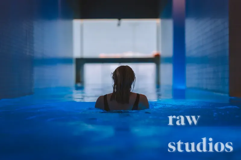 En person med mørkt hår ses bakfra i et blått innendørsbasseng eller spa. Bildet har en rolig stemning med dype blåtoner og dempet belysning.