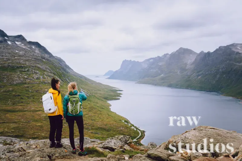 To vandrere med ryggsekker står på en fjellrygg og ser utover en fjord omgitt av grønne fjell og bratte skråninger. Det overskyede lyset gir en rolig stemning i dette vidstrakte landskapet, der fargerike jakker kontrasterer mot de grå og grønne omgivelsene.