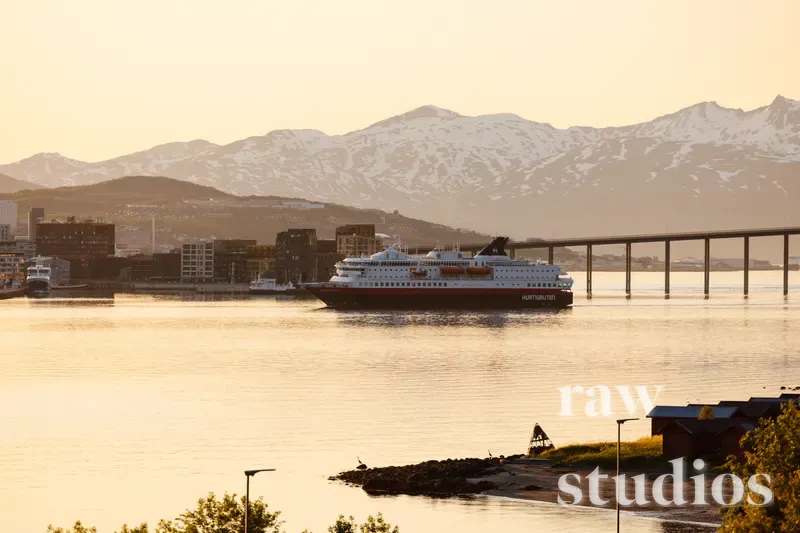 Et stort passasjerskip seiler i en havn med snødekte fjell og en lang bro i bakgrunnen under en gyllen solnedgang. Bildet viser et maritimt landskap med varme farger over vannet og kysten.