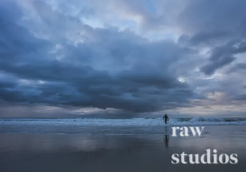 En person går langs strandkanten ved et mørkt hav under en tung, skydekket himmel. Bildet har blå og grå fargetoner som skaper en dramatisk og rå stemning i kystlandskapet.