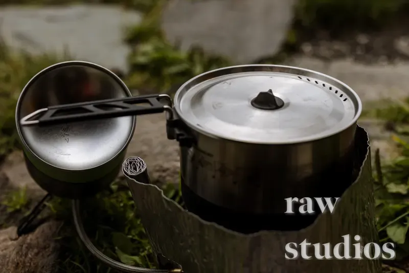En sølvfarget metallgryte med lokk står på en kokeplate omgitt av et vindskjerm i grønn vegetasjon. Bildet viser detaljer fra matlaging under camping eller friluftsliv i naturlig dagslys.